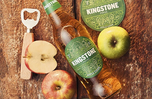 bottle of Kingston press apple cider on wooden planks with bottle opener and apples