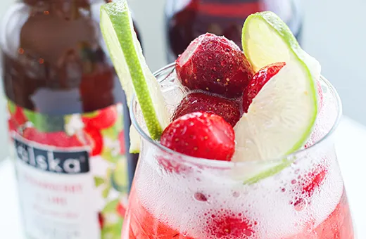 glass of alska strawberry & lime cider with ice and fruit in with bottle in the background